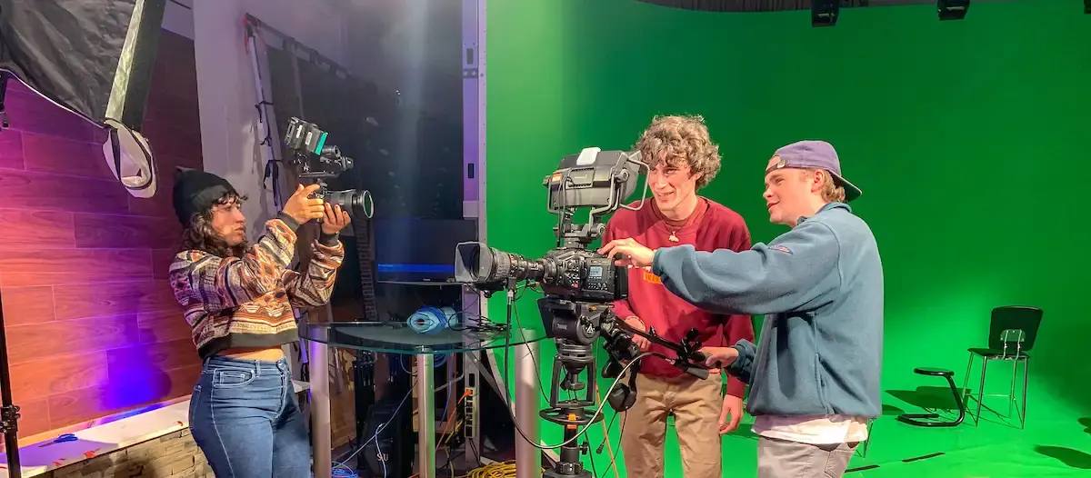 Three students operate professional video cameras in a studio, with one student adjusting a handheld camera and two others collaborating beside a mounted camera in front of a green screen.