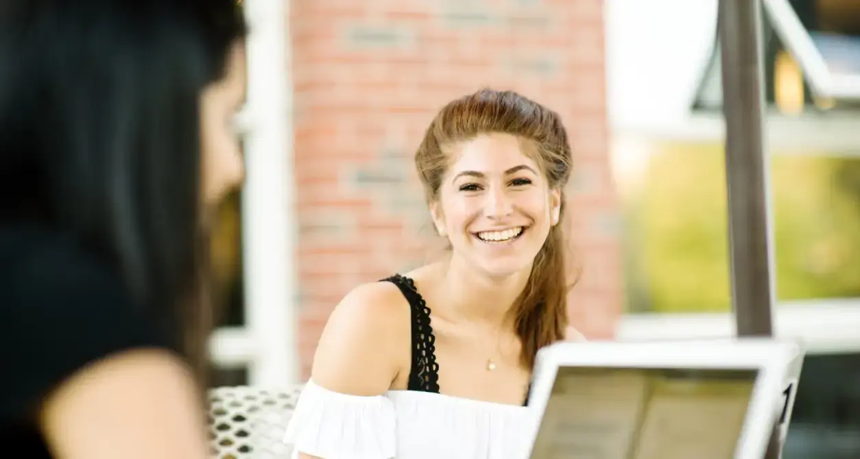 A smiling woman with long, tied-back hair sits indoors, facing another person, with a brick wall and large window in the background.