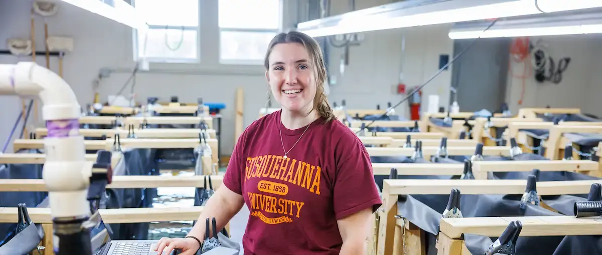 A young woman in a maroon Susquehanna University T-shirt smiles while standing in a laboratory or workshop filled with rows of wooden frames and black tubs.