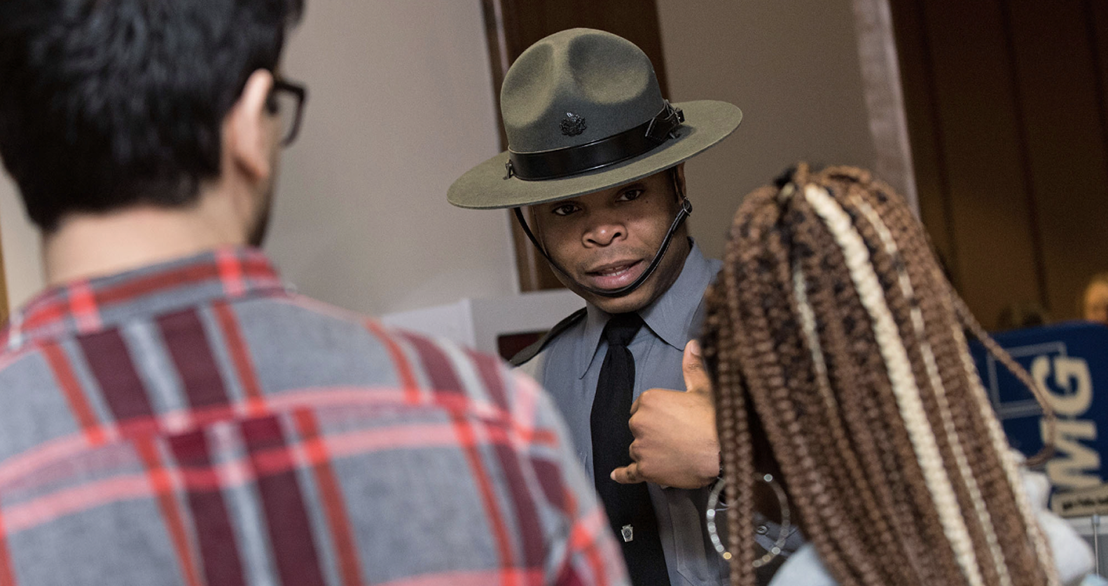 A uniformed officer in a wide-brimmed hat speaks to two individuals, one in a plaid shirt and the other with long braided hair, in an indoor setting.