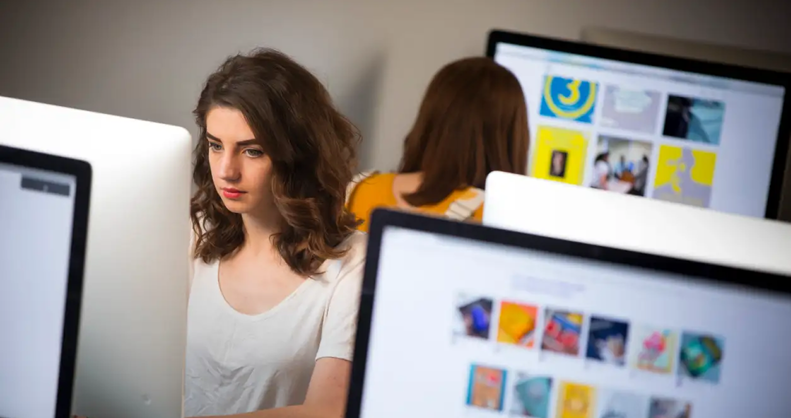 A young woman focuses intently on her computer screen in a modern classroom or studio, with another student visible in the background working on graphic thumbnails.